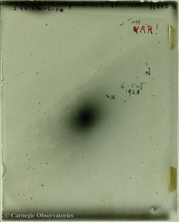 A green-tinted glass plate that is speckled with black spots that are stars, with a large, hazy black oval in the middle. There is some haze surrounding the oval in the shape of a disk. The text written on it has "6 October 1923", a few letter 'N', and one letter 'N' crossed out and replaced with a big red "VAR!"