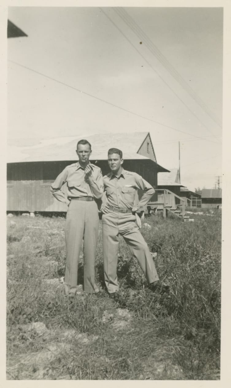 Photo of two men in uniform standing outside military barracks style buildings.