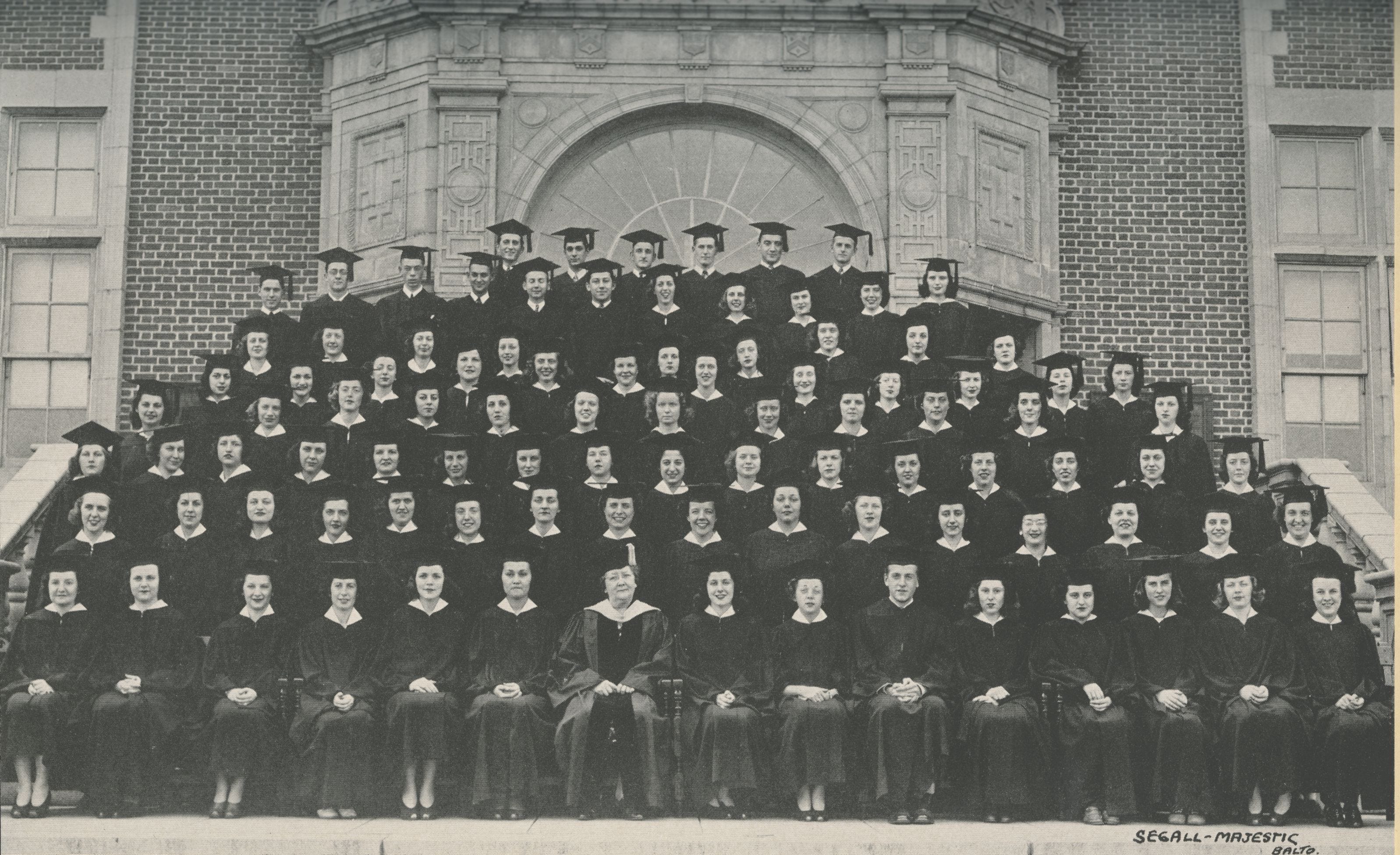 Photo of graduates in caps and gowns arranged on steps in front of Stephens Hall. Dr. Wiedefeld is seated in front and center of the photo dressed in academic regalia